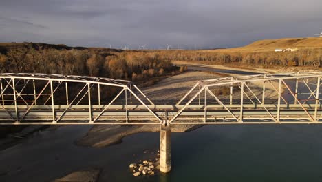 drone approaching and flying over an old steel bridge during sunrise gloom in southern alberta. sustainable wind turbines underneath a dramatic cloudy sky in the background. 4k aerial footage.
