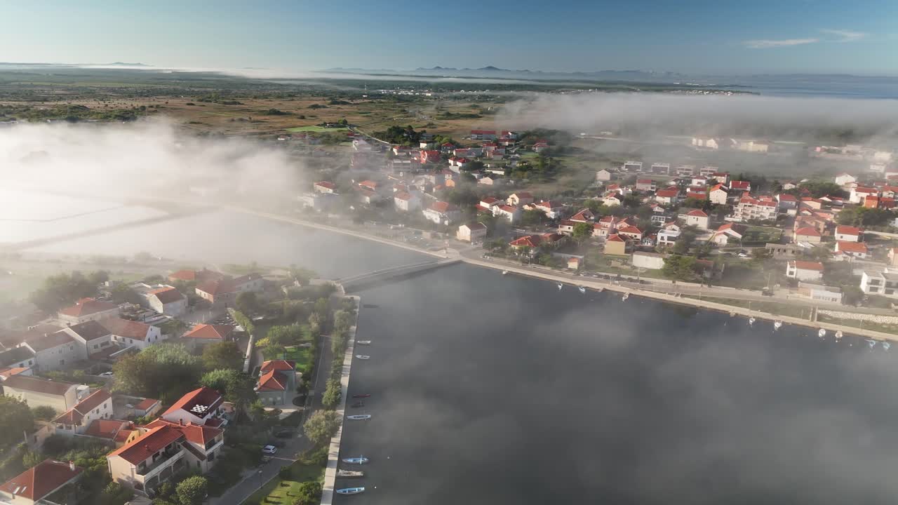 The drone turns and glides above the edge of the island, revealing the narrow sea strait that separates Nin, Croatia from the mainland. Morning mist drifts across the water as the sun breaks through