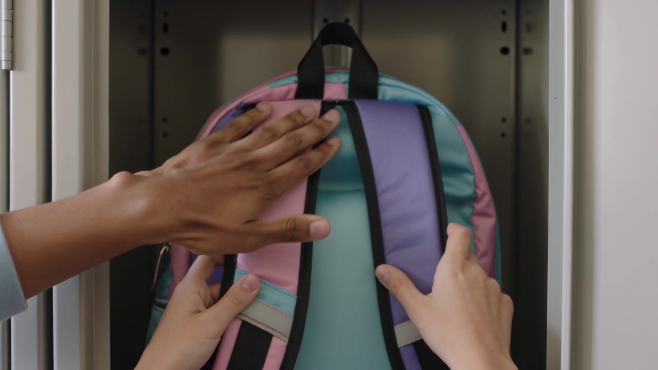 Reaching adult in blue sleeve grasping loop helping child hanging pastel backpack in school locker