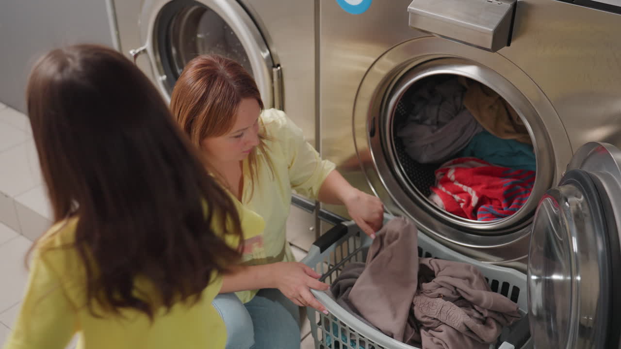 Professional operator kneels by washer removing clothes while little girl watches beside, open door, laundry basket below, stainless machines behind, teamwork moment, family help in modern laundromat