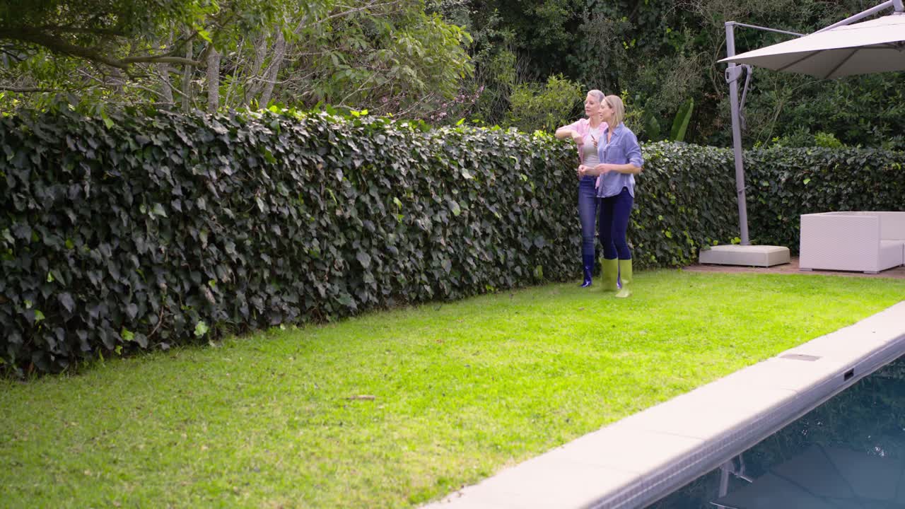 Spotting foliage in garden boots, female friends harvesting and pruning hedge poolside, pointing