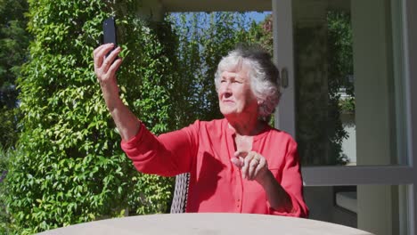 Glückliche-Kaukasische-ältere-Frau,-Die-Am-Tisch-Im-Sonnigen-Garten-Sitzt-Und-Ein-Selfie-Mit-Dem-Smartphone-Macht