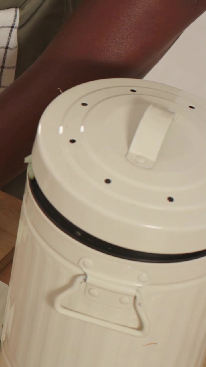 Video verticale: Appoggiando la mano, un cuoco stabilizza un porro su un tagliere per cucinare, con una lattina bianca e un grembiule a quadri.