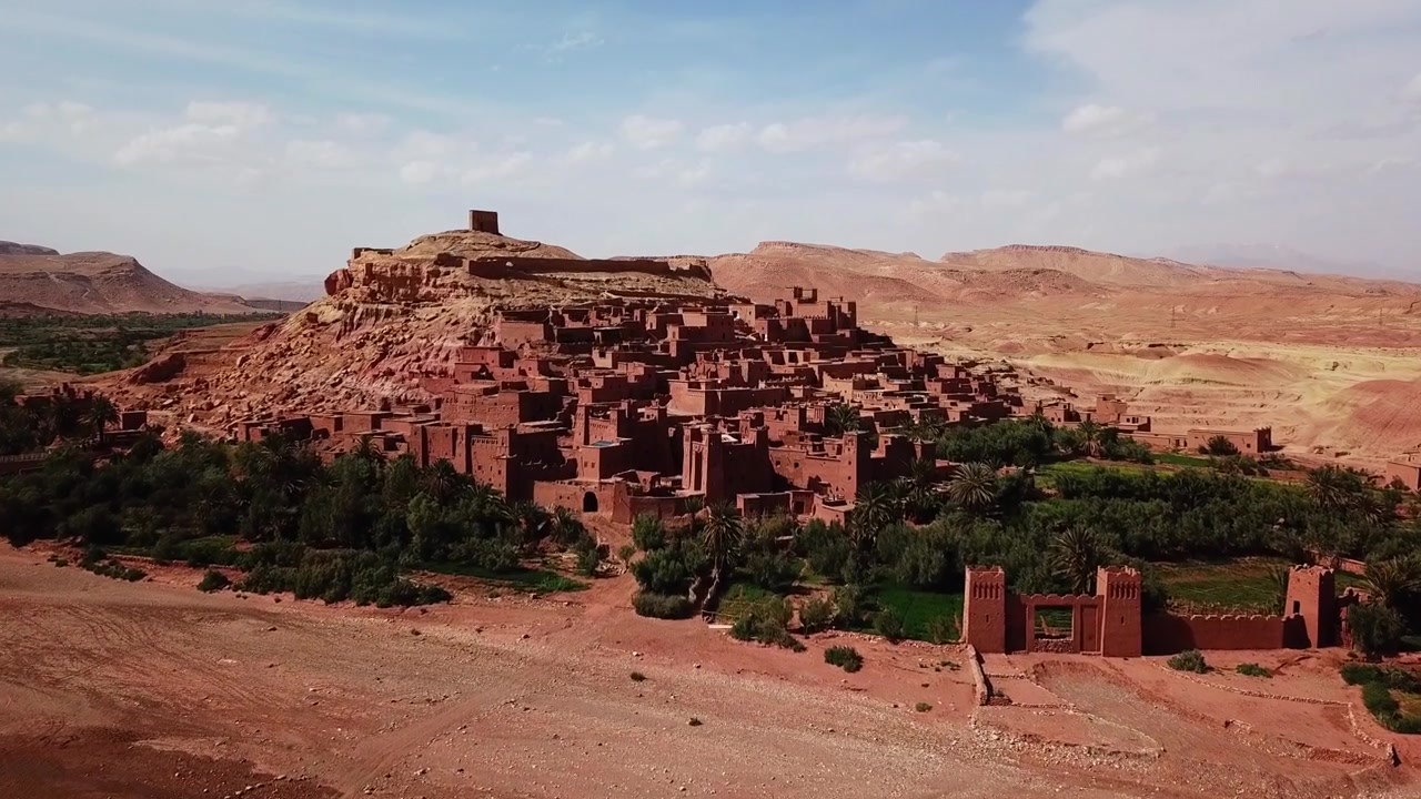Ampia vista cinematografica della kasbah di Ait Benhaddou, circondata da un paesaggio desertico con palme e un'oasi in primo piano. Iconico sito del patrimonio marocchino che fonde architettura e natura, ideale per i viaggi.