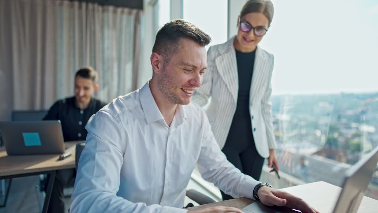 Cheerful smiling colleagues talking in the office. Female stands near her male coworker discussing something and laughing. Male employee sits at desk at backdrop.