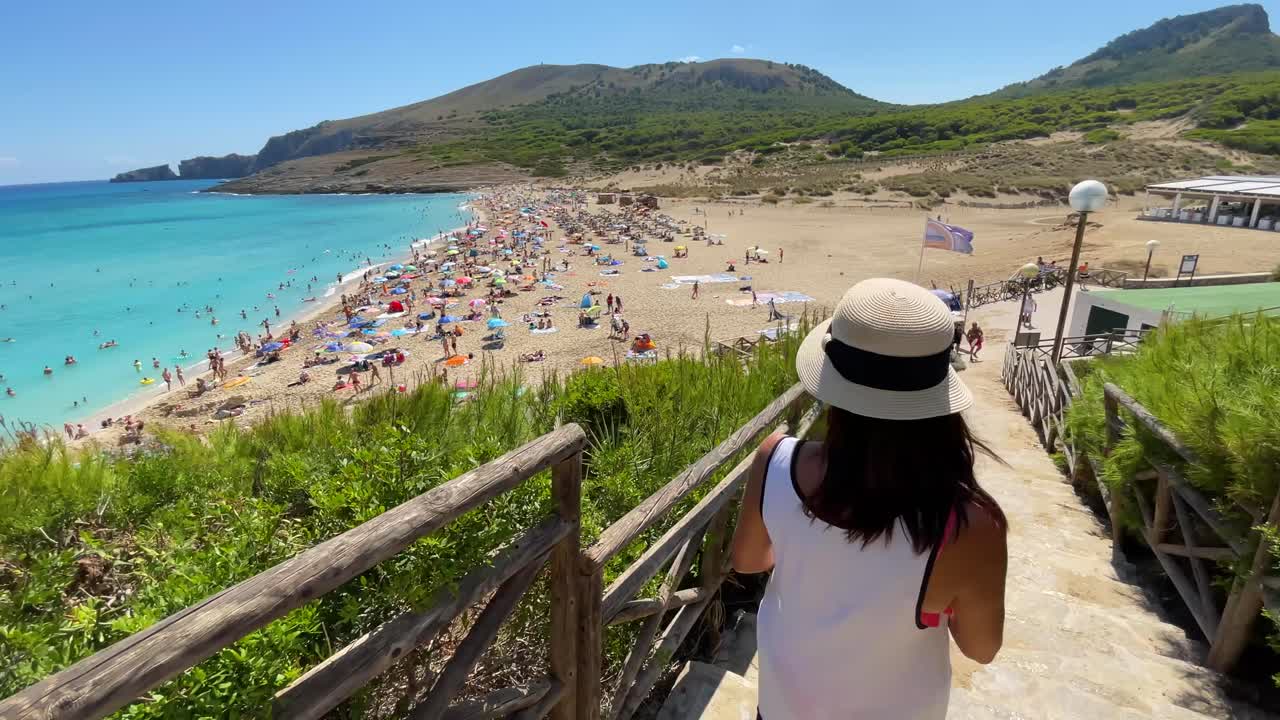 woman with white hat with her back turned down mesquida cove in mallorca balearic islands turquoise blue mediterranean sea, fine white sand beach