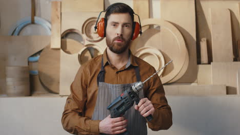 joven carpintero caucásico con auriculares perforando con electrotaladros en el aire en el taller