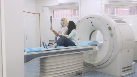 woman doctor radiologist explains the results of ct scanning for young female patient, showing the snapshot with images, observing and analyzing ct scan in modern clinic beside modern ct scanner