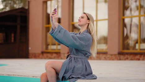 pretty woman in gown is resting in spa hotel with open swimming pool taking selfie by smartphone