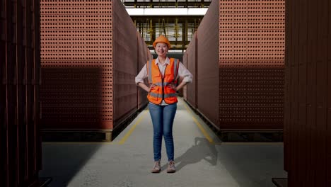 full body of asian female engineer wearing safety helmet looking at camera while standing with arms akimbo with red brick packed in stacks are stored