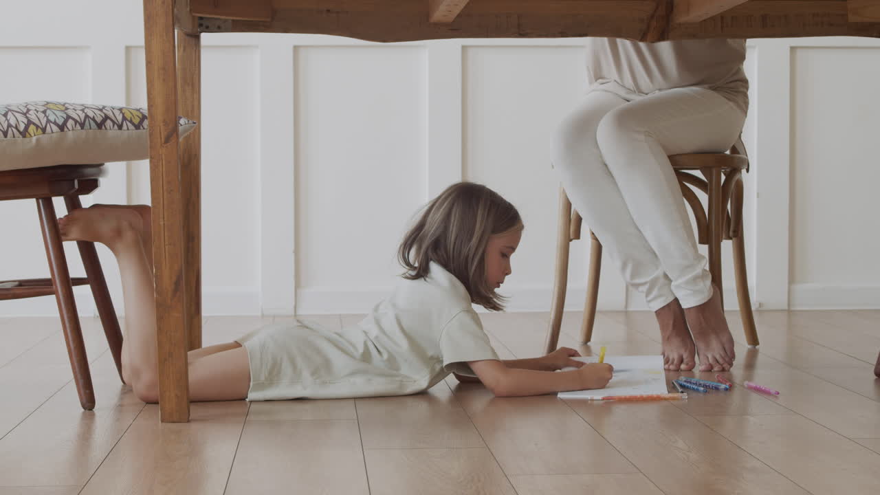 예쁜 금발 소녀는 어머니가 일하는 동안 탁자 아래에 크레용으로 그림을 그리며 주의를 산만하게 합니다.