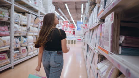 back view of woman in black top and jeans walking near supermarket shelf filled with textile products, adjusting her hair while holding shopping bags, focusing on goods displayed