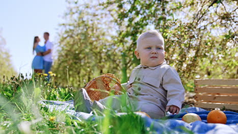 serious cute caucasian blond baby sits on the grass in the garden. couple embracing and kissing at backdrop in blur. low angle view.