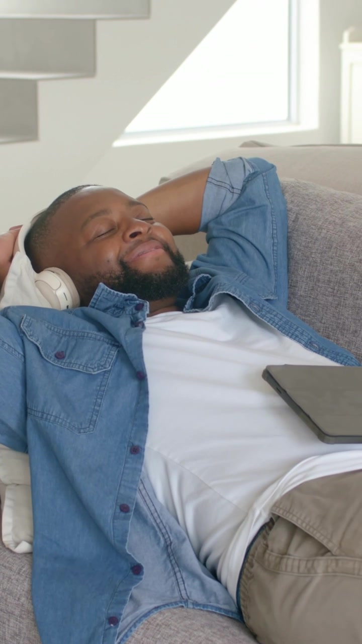 Video verticale: Luce che sorge, uomo in denim che ascolta, sdraiato sul divano, sorridendo con cuffie e tablet.