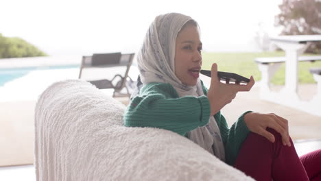 biracial woman in hijab using smartphone on sofa at home with copy space, slow motion