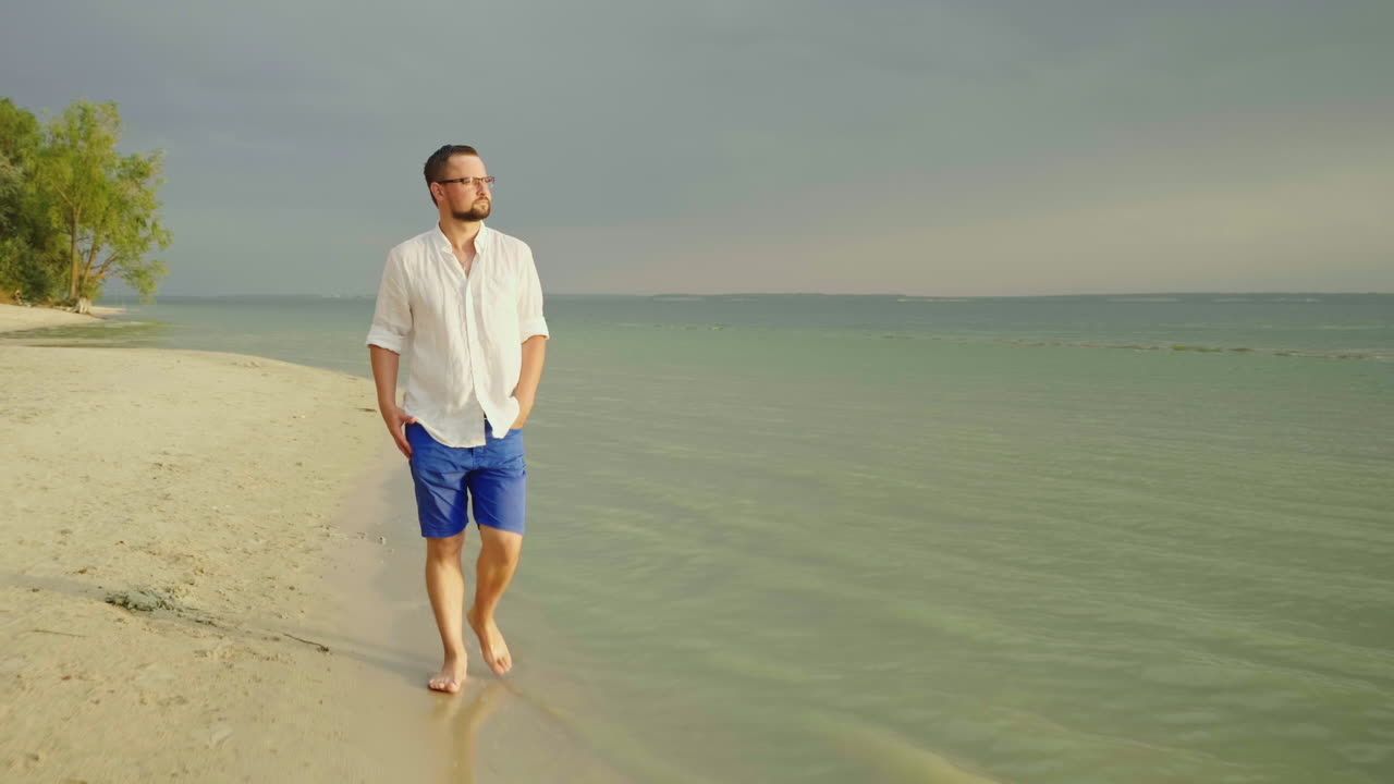 un joven apuesto con ropa ligera y anteojos recorre la playa a lo largo del mar escapando de