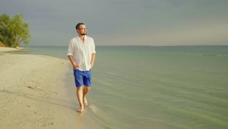 a young well-looking man in light clothes and glasses goes along the beach along the sea escape from