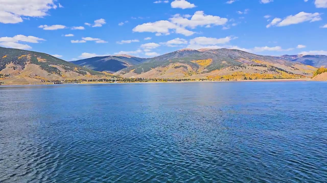 航行中のボートの船首から見た、コロラド州ディロンの広大な山岳湖の風景。深い青色の水、丘の斜面を彩る紅葉、点在するヨット、そして明るい雲。