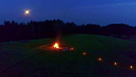 People-warming-up-near-massive-bonfire-at-night,-fly-away-view