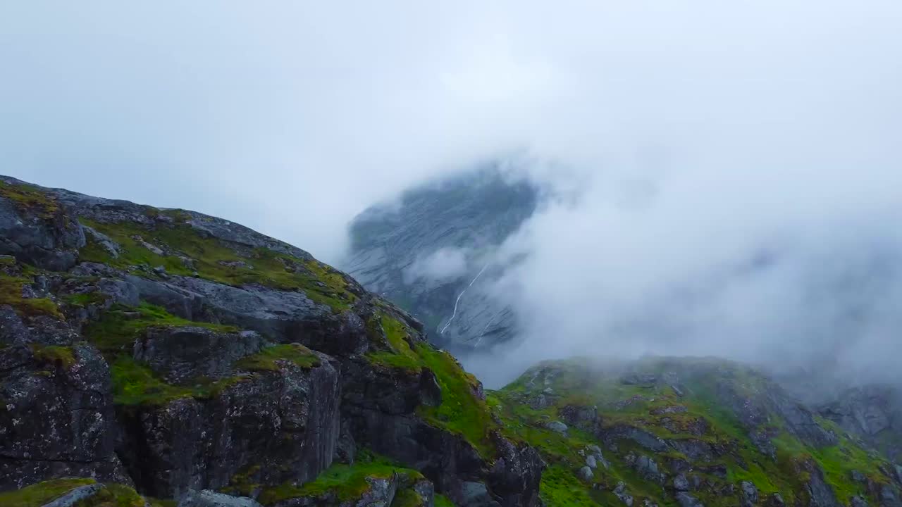 Aerial drone footage gliding forward and flying over Lofoten moss covered and rocky mountains that are between thick white clouds. The cliff edges are steep and sharp, with damp and wet moss on them.