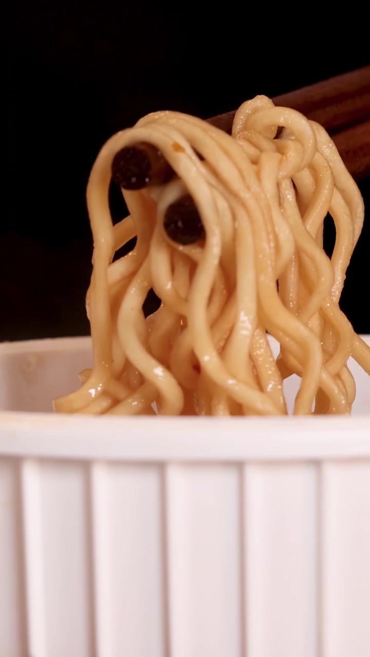 Bacchette afferrano e sollevano noodles istantanei cotti da una tazza bianca a coste su uno sfondo scuro, con illuminazione uniforme e inquadratura ravvicinata
