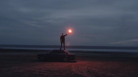 handheld cinematic dark shot of young man standing on the car with red signal burning flare on the beach near the water