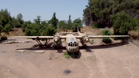 aerial turning and rising drone shot of an old dismantled army plane monument placed on an empty dirt field, surrounded by trees and bushes