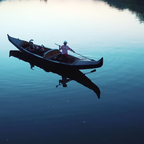 aerial view of a boat floating in the river at sunset. young woman incarnival mask sitting in gondola and a gondolier standing with an oar. camera moves around.