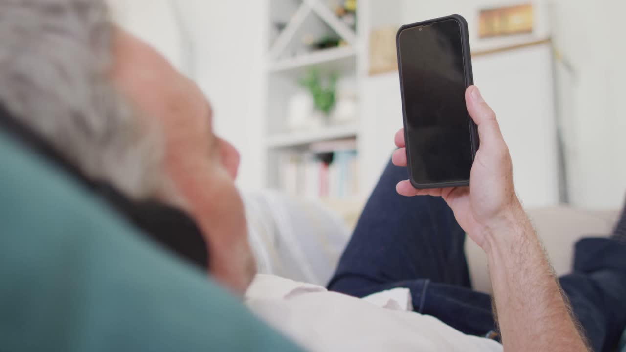 Happy caucasian man lying on sofa in living room, using smartphone