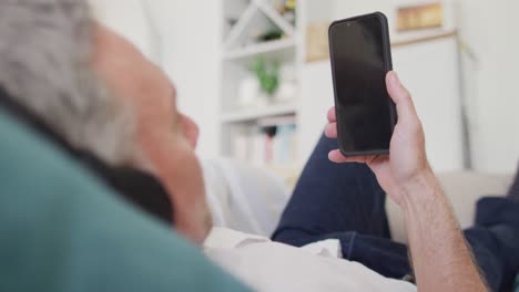 Happy-caucasian-man-lying-on-sofa-in-living-room,-using-smartphone