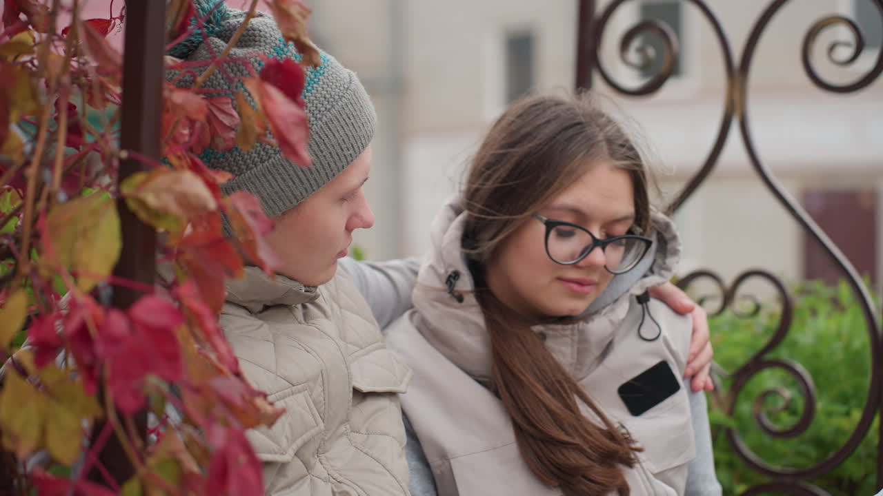 Close up of young couple sitting together on park bench in autumn, man embracing woman gently while she looks at him affectionately, both dressed warmly, surrounded by red leaves