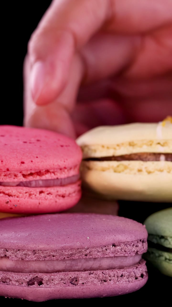 A hand carefully picks assorted colorful macarons from a glossy black surface under bright, even lighting, with close-up camera angles and a clean, minimal background