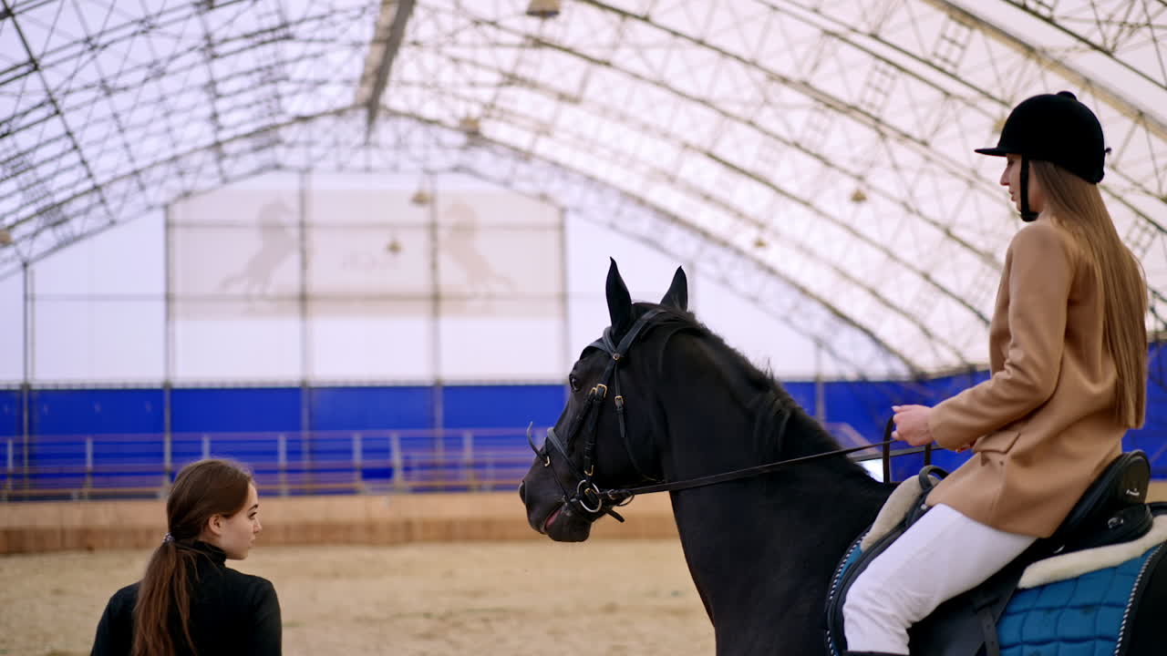 Brunette woman in black helmet riding horse in drill-hall. Female coach holding a horse by the bridle. Manege at backdrop in blur.