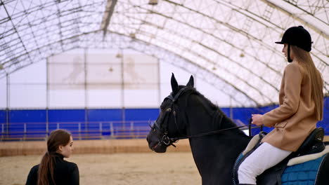 brunette woman in black helmet riding horse in drill-hall. female coach holding a horse by the bridle. manege at backdrop in blur.