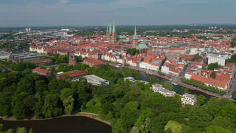 Backwards-reveal-of-green-park-on-island-near-historic-city-centre.-Aerial-panoramic-view-of-town.-Luebeck,-Schleswig-Holstein,-Germany