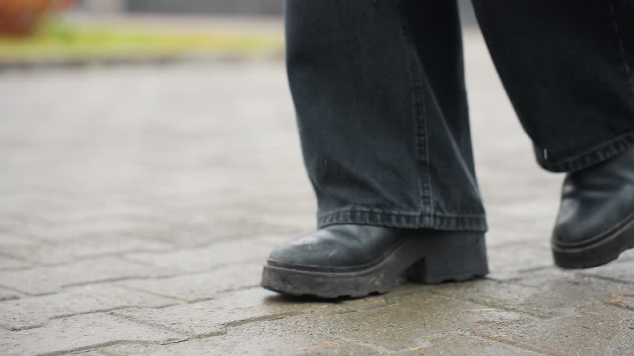 Close up of person's leg wearing black trousers and black boots walking slowly forward on outdoor paved path during calm overcast day, emphasizing footwear motion and moody atmosphere