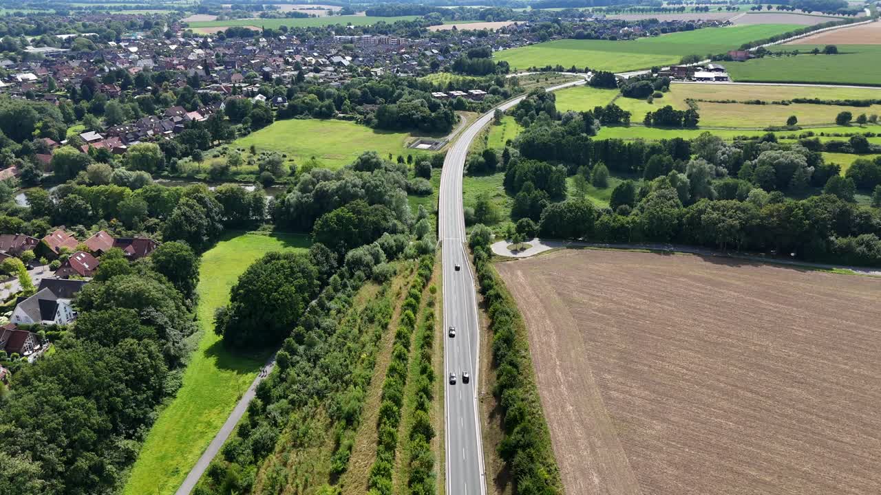 Aerial establishing shot of traffic on American interstate in rural area. Wide shot. Sunny day in scenic area. Green trees and grass fields. Housing area in distance