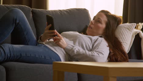 mujer acostada en el sofá en casa harto y aburrido de perder el tiempo usando el teléfono móvil para comprobar el mensaje de las redes sociales y desplazarse en línea 1