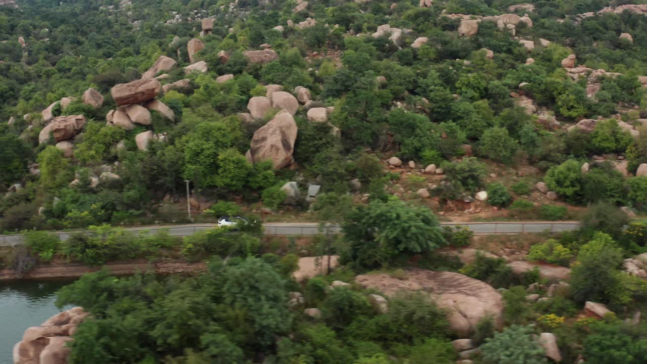 Hvit SUV kjører langs en smal landevei gjennom et frodig, grønt steinete landskap nær Hampi i Karnataka, India