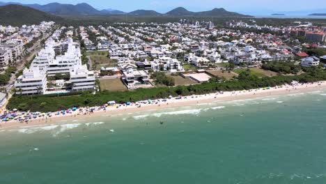 Aerial-drone-hight-altitude-scene-of-tourist-beach-with-many-accommodations-facing-the-sea,-flight-buildings-and-houses-with-people-having-fun-on-the-beach-in-Florinópolis-Jurere-Internacional