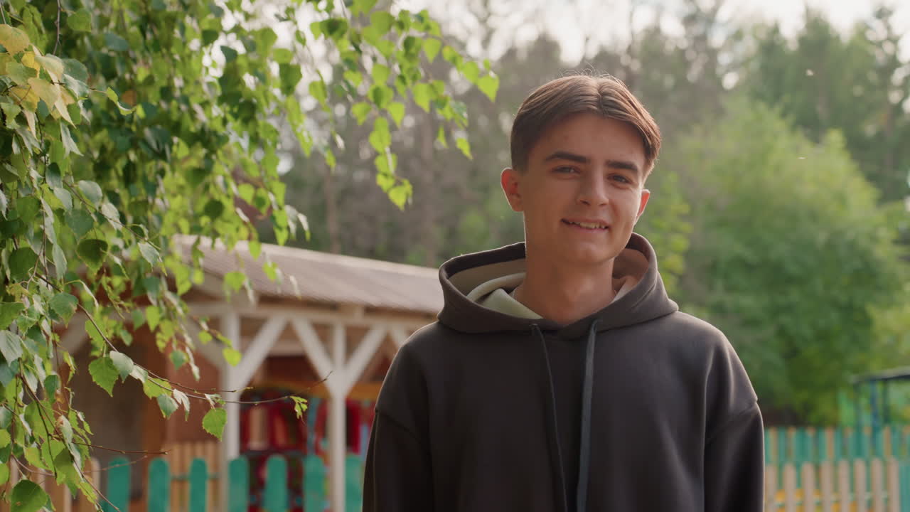 White teenage boy smiling near gazebo and playground, relaxed portrait with bright greenery and warm sunlight. Cheerful student chatting with friends, campus volunteer greeting passerby, aspiring