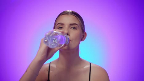 Thirsty-woman-drinks-water-from-a-transparent-plastic-bottle,-purple-background