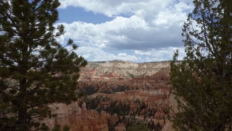 tilt down shot revealing a beautiful southern utah desert valley landscape framed by two pine trees with large hoodoo formations from erosion surrounded by greenery on a cloudy sunny summer day