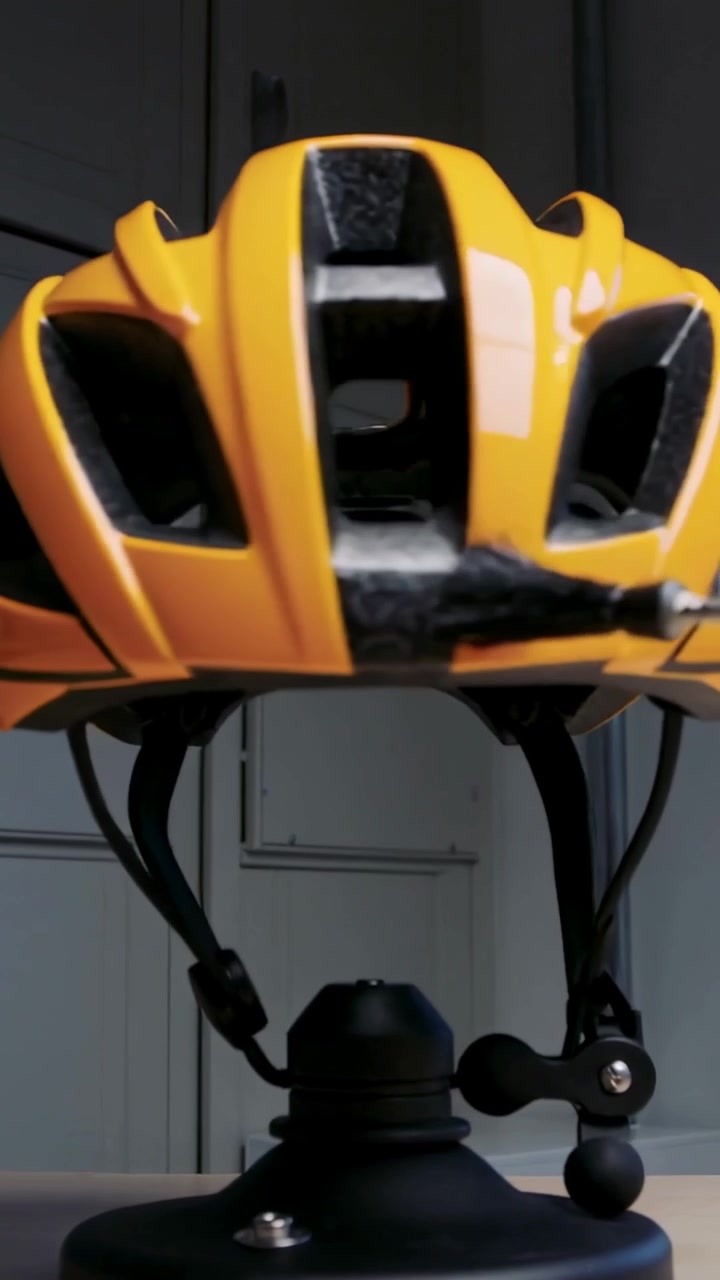 Vídeo vertical: Capacete de bicicleta amarelo girando em suporte giratório preto em uma bancada, mostrando aberturas e encaixe.