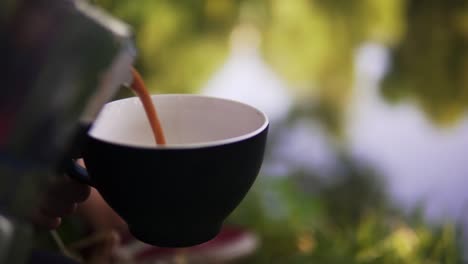 beautiful shot of a person filling coffee into a cup at a camping trip with the trees reflecting in the water