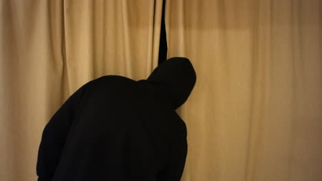 a man in a black hood is suspicious and in opening the curtains to spy outside