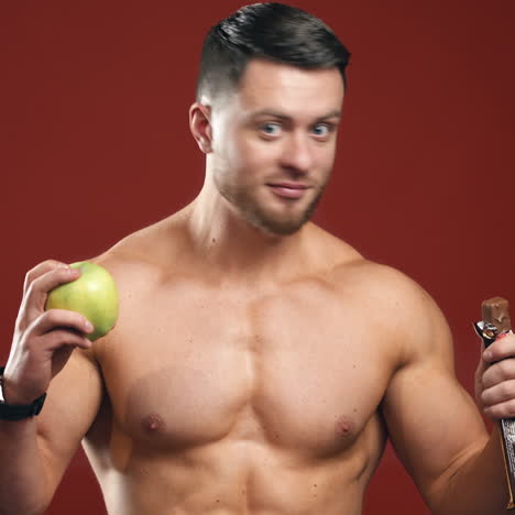 portrait of a athletic man choosing between a fresh apple and a chocolate. healthy food. dieting concept. half lenght video.