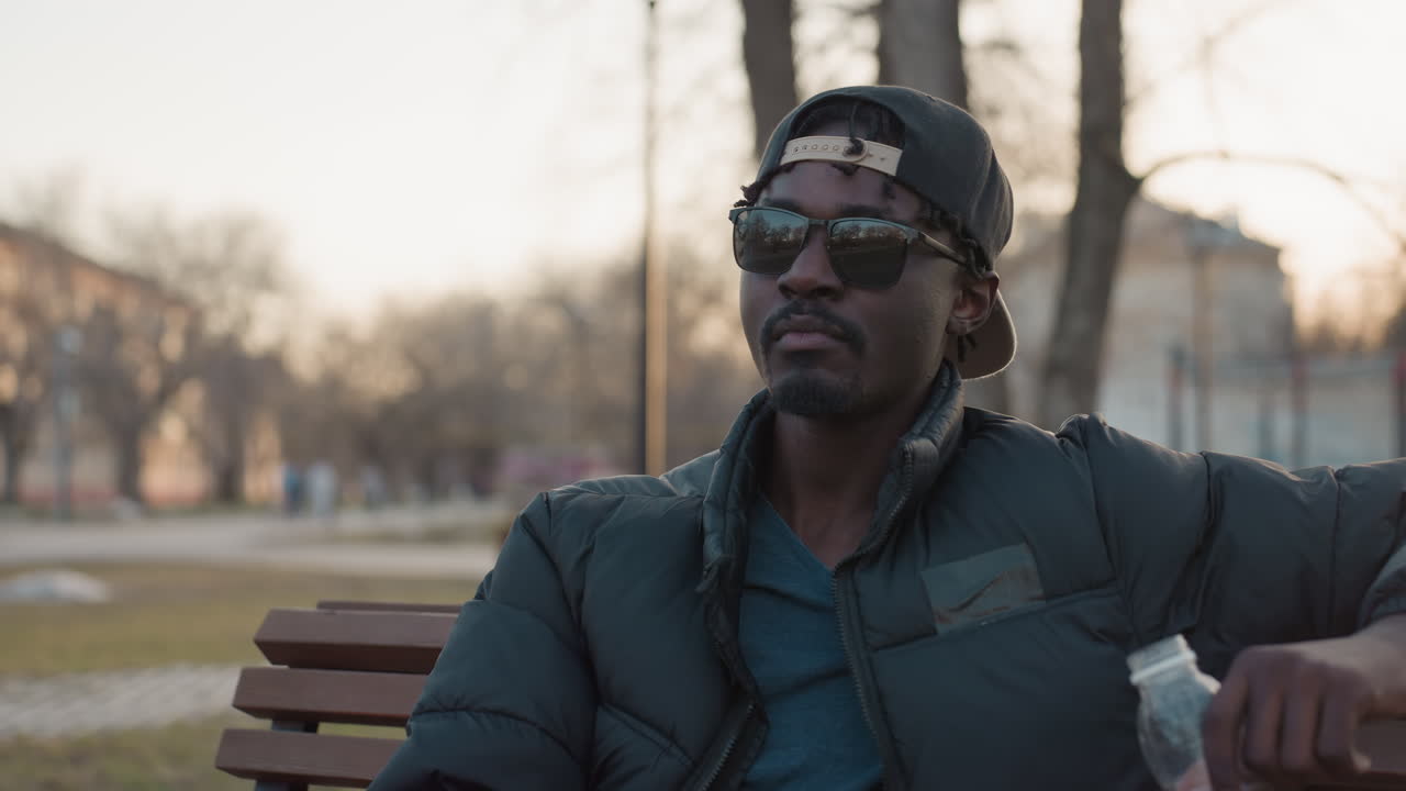Young man in sunglasses sits on park bench sipping juice from bottle, cap worn backward, relaxed posture, sunset glow, blurred people and trees behind, quiet urban leisure moment with calm mood
