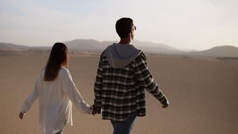 happy couple holding hands walking in romantic relationship under sun and blue sky in desert. two young lovers walking by desert sand in casual clothes and smiling