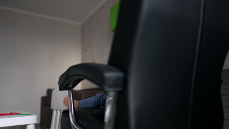 adorable caucasian toddler sits in the rotating office chair. cute toddler is focused on smartphone. low angle view.